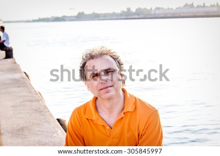 Classy  40 years old sportsman with three-day beard and salt and pepper hair wearing an orange polo shirt while he is sitting on the pier and resting