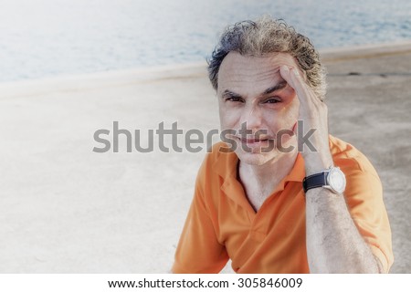Classy  40 years old sportsman with three-day beard and salt and pepper hair wearing an orange polo shirt while he is sitting on a bench on the pier and holding his head: is it headache?