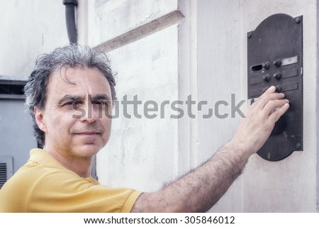 Classy  40 years old sportsman with three-day beard and salt and pepper hair wearing a yellow polo shirt while he is ringing a bell of an apartment building in residential neighborhood