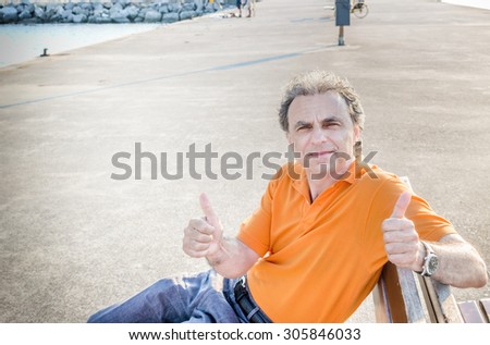 Classy  40 years old sportsman with three-day beard and salt and pepper hair wearing an orange polo shirt while he is sitting on a bench on the pier and showing thumbs up