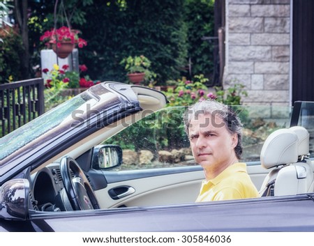 Side view of classy  40 years old sportsman with three-day beard and salt and pepper hair wearing a yellow polo shirt while he is driving a dark brown car in residential neighborhood