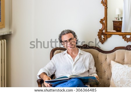 fascinating forties man dressed in a refined yet sporty manner is wearing glasses and watching a coffee table photo book on an upholstered sofa in a bright and luxurious living room