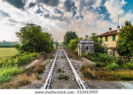 Rails of a country railroad crossing  under a cloudy sky in Summer in Italy