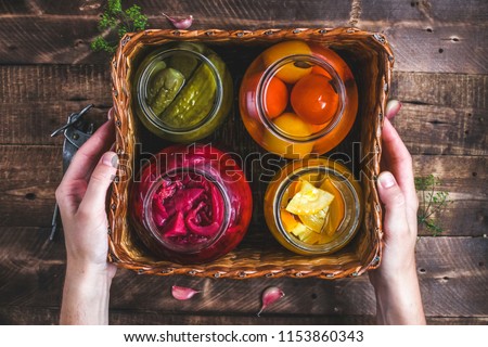 Homemade glass jars of pickled fresh cucumbers, juicy tomatoes, sweet zucchini in a wicker box on a wooden background. Stock of food. Autumn concept. Top view