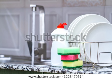 Different clean plates in dish drying rack, dish sponges and dishwashing detergent on the table on kitchen counter. Washing dirty dishes