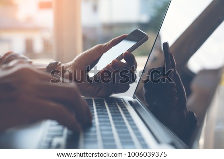 Close up of casual man or freelancer working on laptop computer and holding mobile smart phone with reflection on blank screen, working from home or casual business concept.