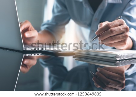 Business planning, E learning concept. Casual business man writing on notebook and working on laptop computer in modern office. Man studying online course via laptop and lecture on notepad.
