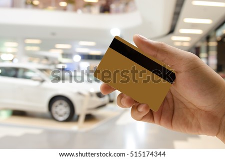 Man hand holding empty golden credit or debit card, member card, vip card with blurred background of new cars in showroom, or motor show event, credit card payment, car insurance concept.