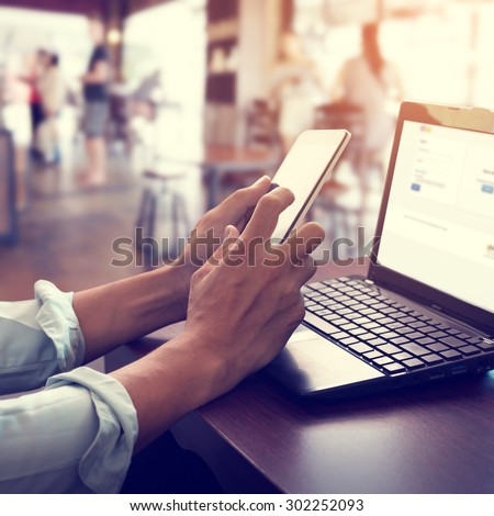 Side view shot of young business man working on his laptop and using smart phone sitting at wooden table in a coffee shop with retro filter effect