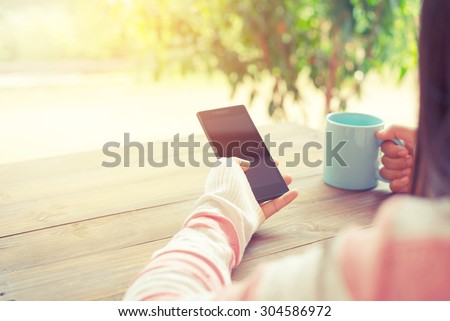 Rear view of female hand holding smart phone with hot cup of coffee on wood table. Photo in vintage pastel color image style.