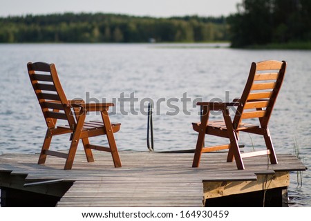 A lone wooden chairs sitting on the dock with a lake and cottages across