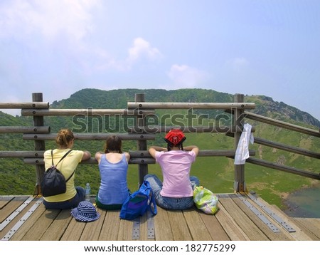 three girls sitting on a wooden observation deck facing mountains on Jeju Island, South Korea