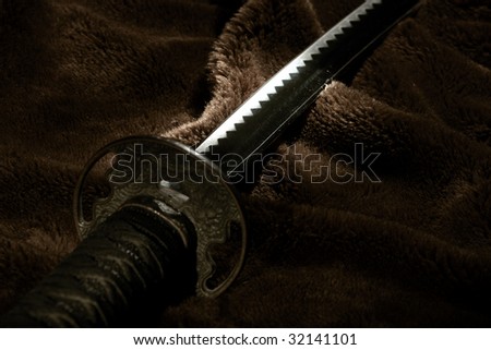 Samurai sword laying on brown fabric. Lighting hitting the blade with focus on the blade just above the hilt.