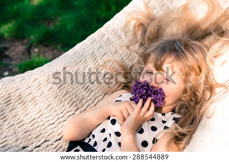 beautiful blond girl with long blond hair lying in white hammock and smelling lilac flower
