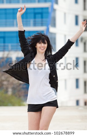 Attractive brunette in black on open air. Young woman in black shorts and jacket runs along the street with arms raised