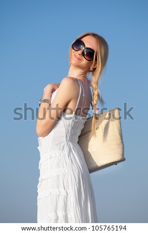 Attractive young woman in white dress and sunglasses posing on background of sky. Girl with bag looking over shoulder
