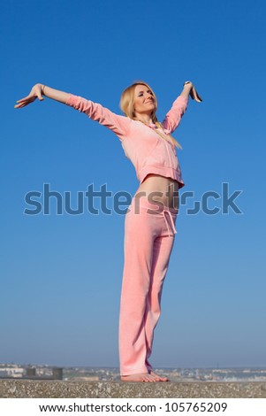 Attractive young woman in pink sportswear posing on background of sky. Barefoot girl with hands raised on open air