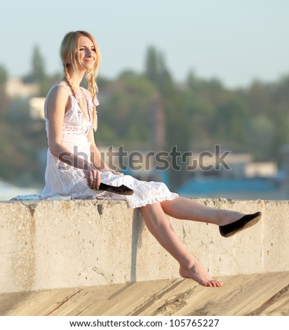 Attractive young woman in white sundress puts on shoes on open air. Girl with pigtails puts on shoes looks away smiling