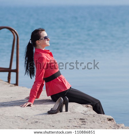 Young woman on seashore. Brunette in a red blouse sitting on the pier at the sea