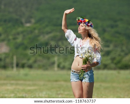 Ukrainian girl in field. Attractive blond woman in ukrainian wreath and blouse with ukrainian embroidery posing on open air taking bouquet of wild chamomile