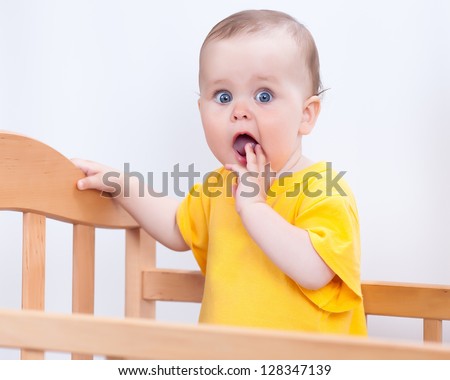 Charming kid in the crib. Baby girl in yellow looking at camera through fence