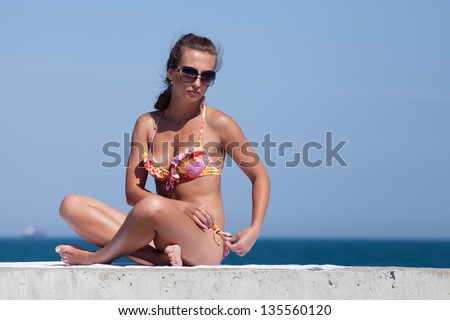 Young woman in bikini at the sea. Attractive woman in swimwear sitting on pier and correcting her swimming trunks