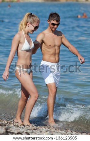 Attractive couple at the sea. Young man and woman in white swimwear and sunglasses walking along seashore in day time