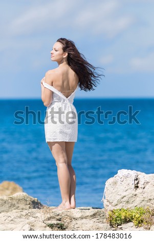 Young woman at the sea. Curly girl in white standing on rocky seashore looking away