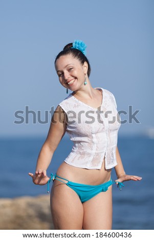 Girl at the sea. Young woman in white blouse and blue swimming trunks posing on background of sea looking at camera smiling