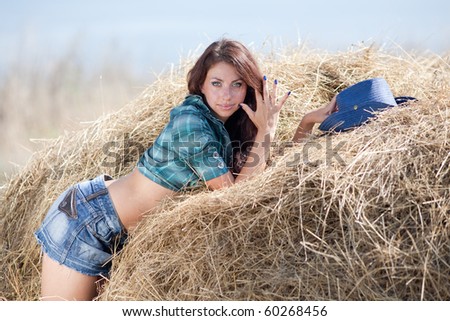 Attractive brunette looks at camera outdoors. Young woman in costume of cowboy lies on haystack