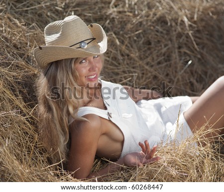 Portrait of blond girl in white dress and hat. Young woman in costume of cowboy lies on haystack