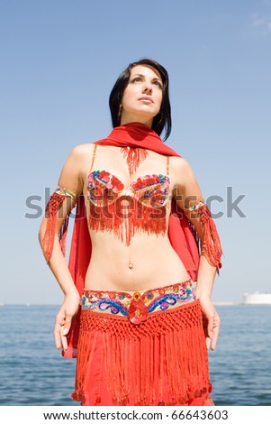 Attractive brunette in red costume for oriental dancing on the seashore. Young woman is dancing belly dance outdoors