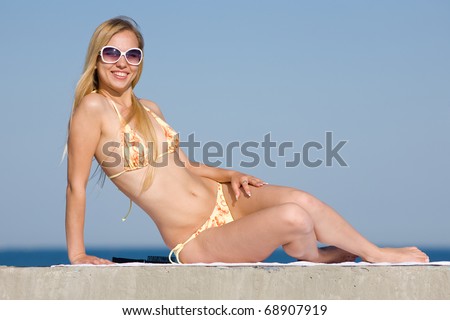 Blonde in bikini and sunglasses at the sea. Attractive young woman in bikini sitting on the beach