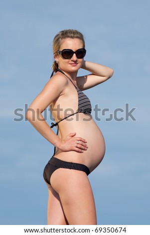 Pregnant woman in swimwear outdoors. Attractive expectant mother in bikini looks through sunglasses at camera