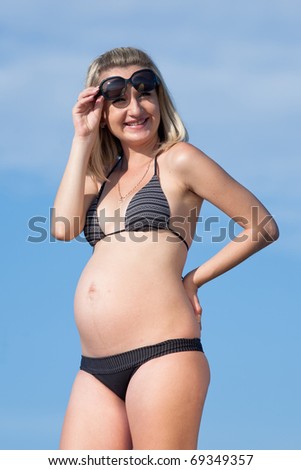 Pregnant woman in swimwear outdoors. Attractive expectant mother in bikini looking from under sunglasses at camera smiling