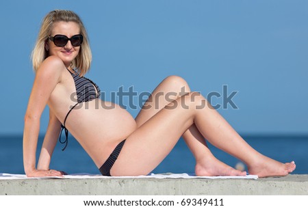 Pregnant woman in swimwear outdoors. Attractive expectant mother in bikini sits and looks through sunglasses at camera