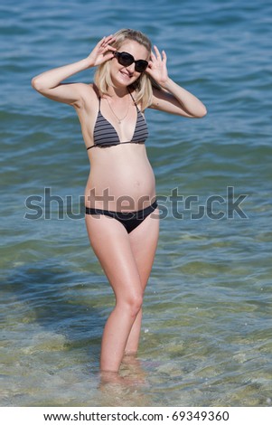 Pregnant woman in swimwear at the sea. Attractive expectant mother in bikini looks through sunglasses at camera