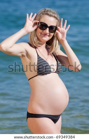 Pregnant woman in swimwear at the sea. Attractive expectant mother in bikini looks through sunglasses at camera