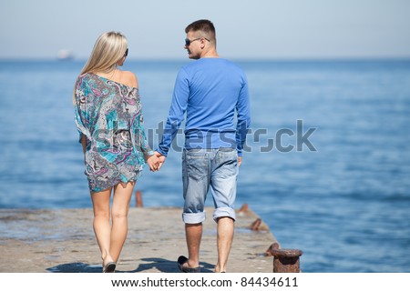 Attractive couple walking along pier. Young man and young woman in full-back position
