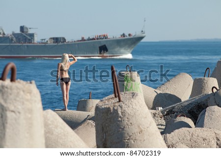 Attractive blonde in black bikini observes navy ship. Attractive young woman at the sea