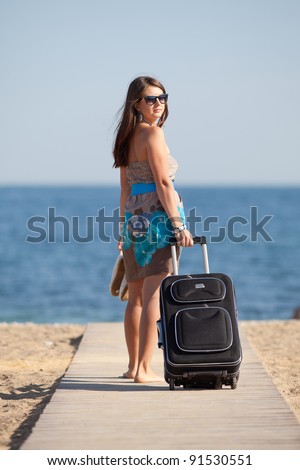 Attractive young woman with suitcase on the beach. Barefoot young woman in dress and sunglasses with rolling suitcase walking along the beach