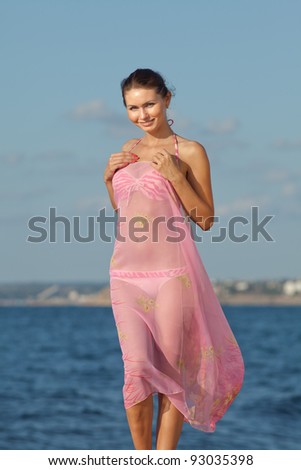 Girl at the sea. Attractive young woman is posing on background of sea. Lady in pink sarong with arms raised looking at camera smiling