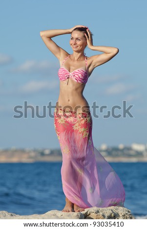 Girl at the sea. Attractive young woman is posing on background of sea. Lady in pink sarong with arms raised on the beach