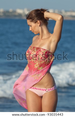 Girl at the sea. Attractive young woman is posing on background of sea. Lady in pink sarong with arm raised on the beach