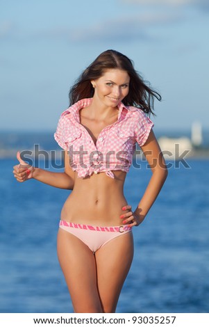 Girl at the sea. Attractive young woman showing thumb up on background of sea. Lady in pink shirt showing Ok looking at camera smiling