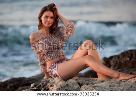Lady at the sea. Attractive young woman in shorts and blouse on background of sea. Barefoot girl sitting on rock