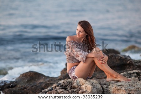 Lady at the sea. Attractive young woman in shorts and blouse on background of sea. Barefoot girl sitting on rock