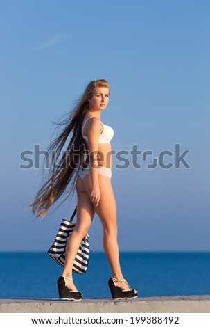Young woman in white lingerie with beach bag walking along seashore looking at camera smiling