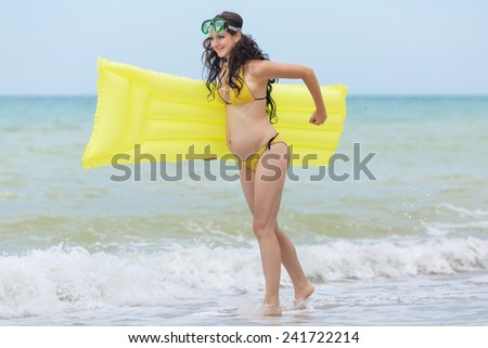 Expectant mother with pool raft and diving mask playing at the sea. Pregnant woman in yellow bikini playing on the beach