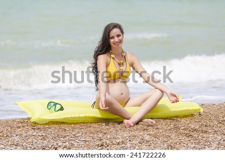 Expectant mother sits on pool raft looking at camera smiling. Pregnant woman in yellow bikini on pebble beach